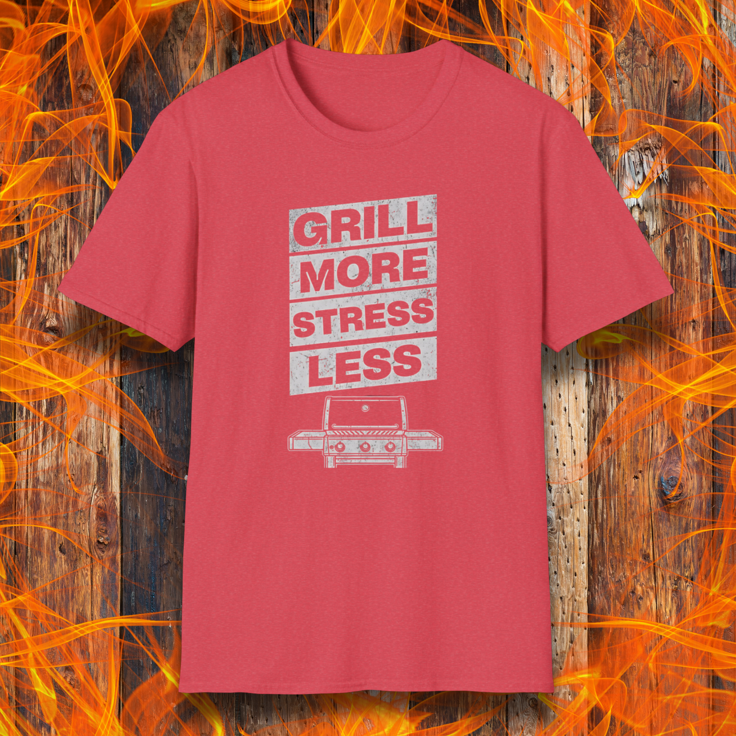 Red t-shirt with 'Grill More Stress Less' text and graphic on a wooden background with flames.