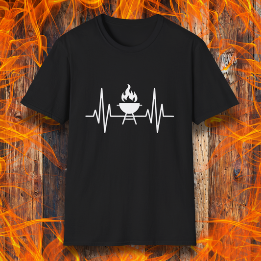 Black T-shirt featuring a minimalist black design of a heartbeat line that transitions into a flaming charcoal grill in the center. The design symbolizes a passion for BBQ, integrating a clean and modern aesthetic. The background consists of a rustic wooden surface with vivid orange flames, enhancing the fiery and grilling-focused theme.