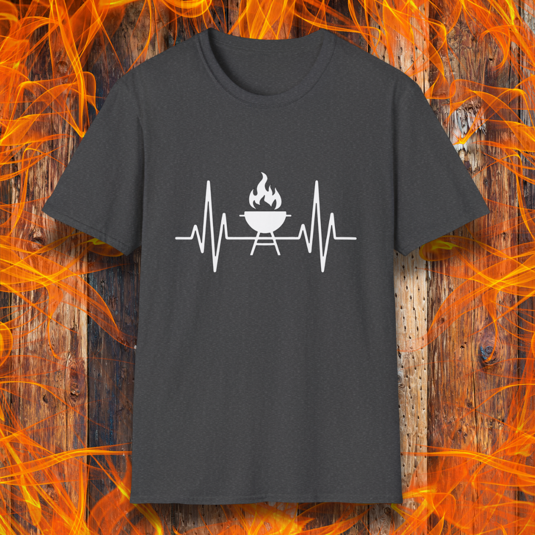 Dark Heather T-shirt featuring a minimalist black design of a heartbeat line that transitions into a flaming charcoal grill in the center. The design symbolizes a passion for BBQ, integrating a clean and modern aesthetic. The background consists of a rustic wooden surface with vivid orange flames, enhancing the fiery and grilling-focused theme.