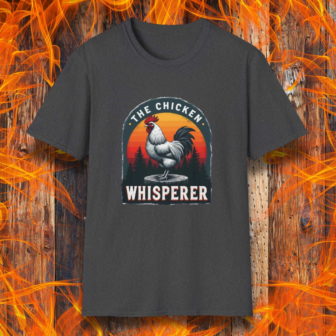 Dark Heather T-shirt featuring a bold design with a proud rooster standing on a wooden stump. The rooster is framed by an arched emblem with a scenic sunset gradient in orange, red, and yellow hues, along with a silhouette of pine trees in the background. The text "THE CHICKEN WHISPERER" is displayed prominently, giving the design a humorous and rustic feel, perfect for backyard BBQ enthusiasts and poultry lovers.