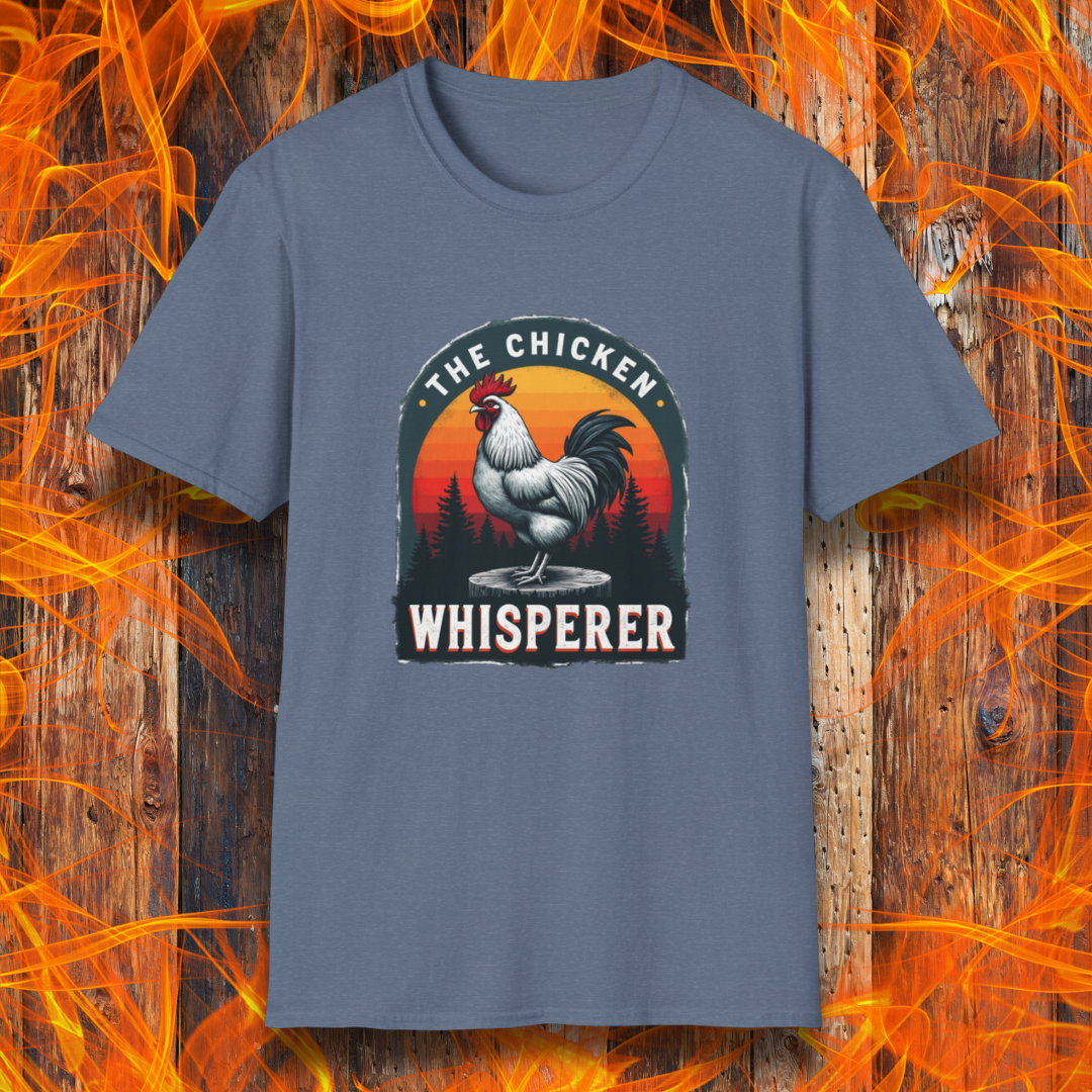 Heather Indigo T-shirt featuring a bold design with a proud rooster standing on a wooden stump. The rooster is framed by an arched emblem with a scenic sunset gradient in orange, red, and yellow hues, along with a silhouette of pine trees in the background. The text "THE CHICKEN WHISPERER" is displayed prominently, giving the design a humorous and rustic feel, perfect for backyard BBQ enthusiasts and poultry lovers.
