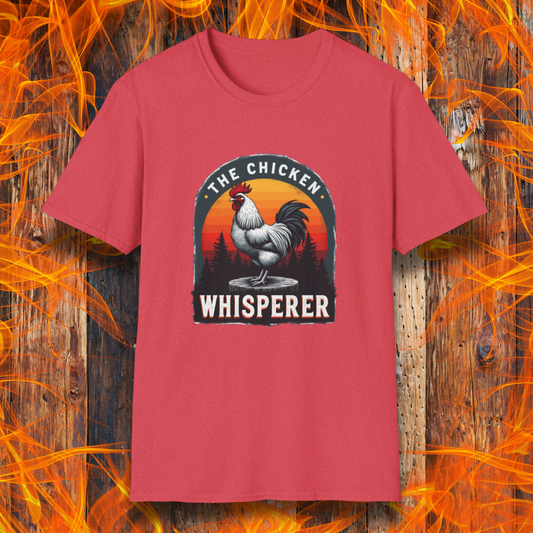 Heather Red T-shirt featuring a bold design with a proud rooster standing on a wooden stump. The rooster is framed by an arched emblem with a scenic sunset gradient in orange, red, and yellow hues, along with a silhouette of pine trees in the background. The text "THE CHICKEN WHISPERER" is displayed prominently, giving the design a humorous and rustic feel, perfect for backyard BBQ enthusiasts and poultry lovers.