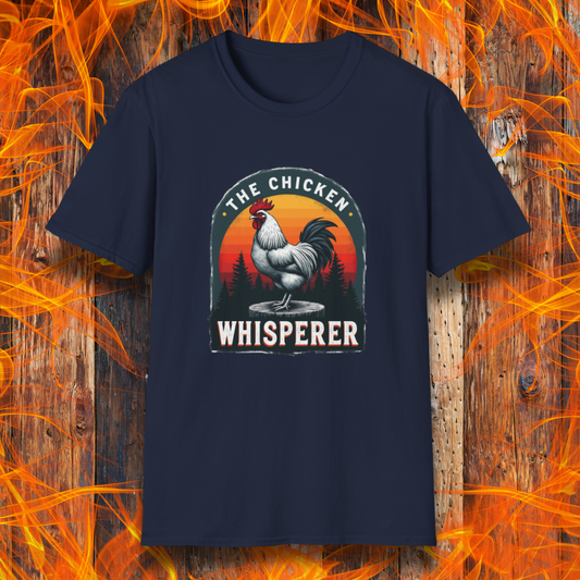 Navy T-shirt featuring a bold design with a proud rooster standing on a wooden stump. The rooster is framed by an arched emblem with a scenic sunset gradient in orange, red, and yellow hues, along with a silhouette of pine trees in the background. The text "THE CHICKEN WHISPERER" is displayed prominently, giving the design a humorous and rustic feel, perfect for backyard BBQ enthusiasts and poultry lovers.