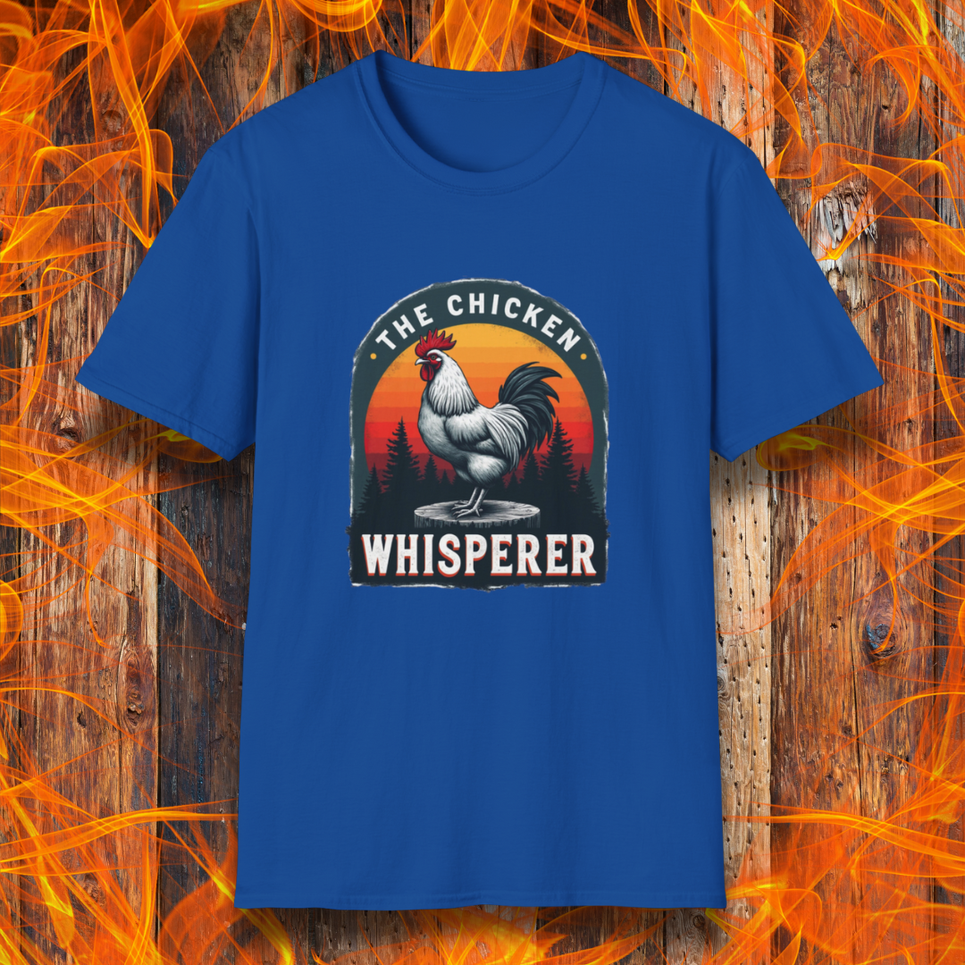 Royal Blue T-shirt featuring a bold design with a proud rooster standing on a wooden stump. The rooster is framed by an arched emblem with a scenic sunset gradient in orange, red, and yellow hues, along with a silhouette of pine trees in the background. The text "THE CHICKEN WHISPERER" is displayed prominently, giving the design a humorous and rustic feel, perfect for backyard BBQ enthusiasts and poultry lovers.