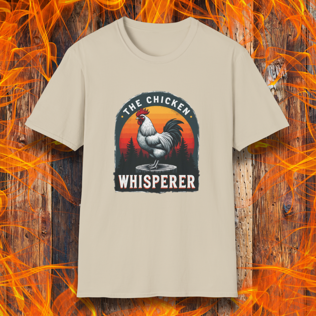 Sand T-shirt featuring a bold design with a proud rooster standing on a wooden stump. The rooster is framed by an arched emblem with a scenic sunset gradient in orange, red, and yellow hues, along with a silhouette of pine trees in the background. The text "THE CHICKEN WHISPERER" is displayed prominently, giving the design a humorous and rustic feel, perfect for backyard BBQ enthusiasts and poultry lovers.