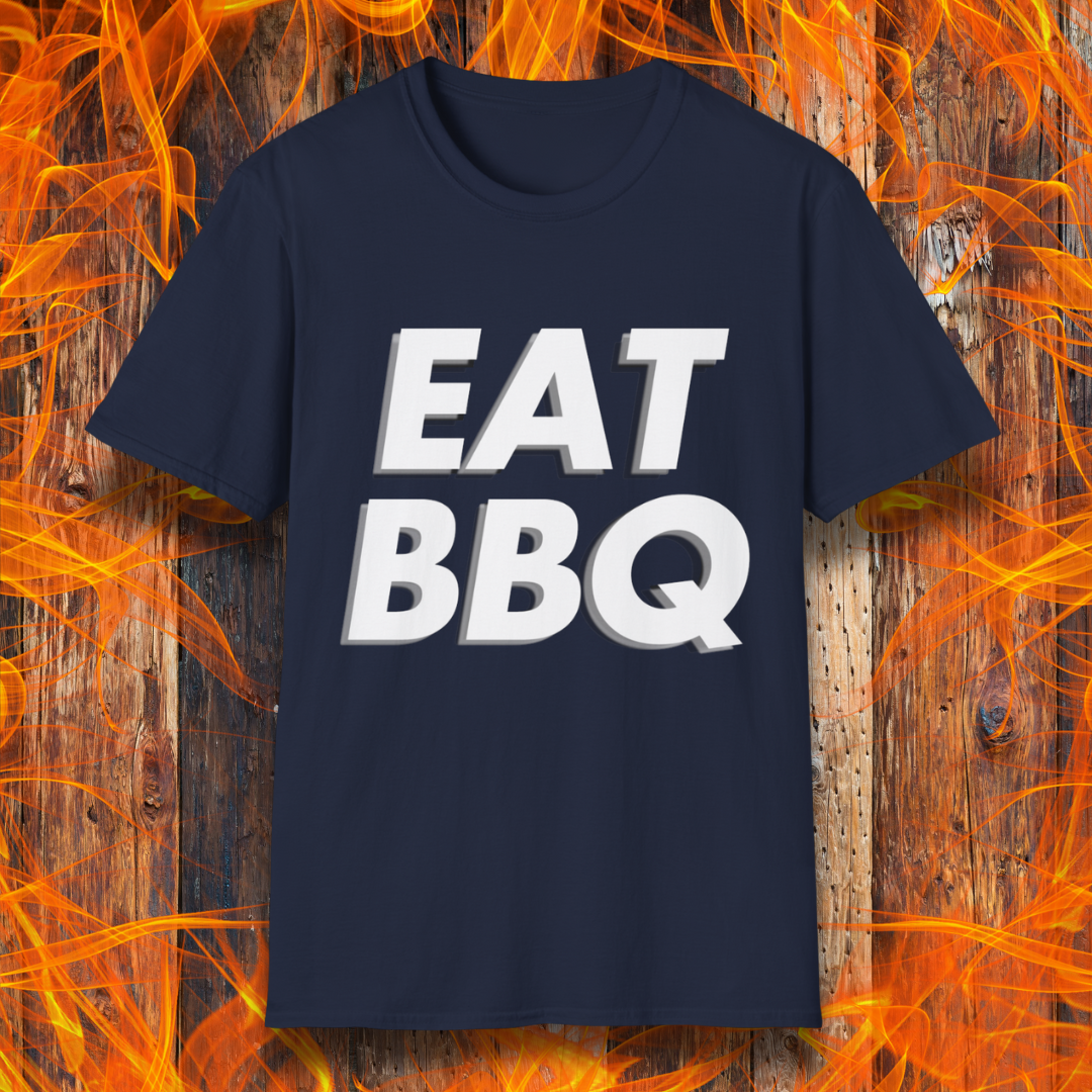 Navy T-shirt featuring bold, blocky white text that reads "EAT BBQ" with a 3D effect. The shirt is displayed against a fiery wooden background, emphasizing the BBQ theme.