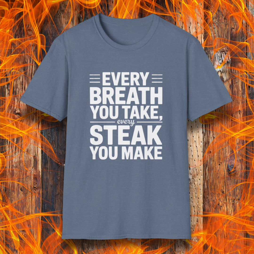 Heather Indigo T-shirt with bold white text that reads "Every Breath You Take, Every Steak You Make," displayed over a rustic wood and flame background — a humorous grilling shirt perfect for steak fans.