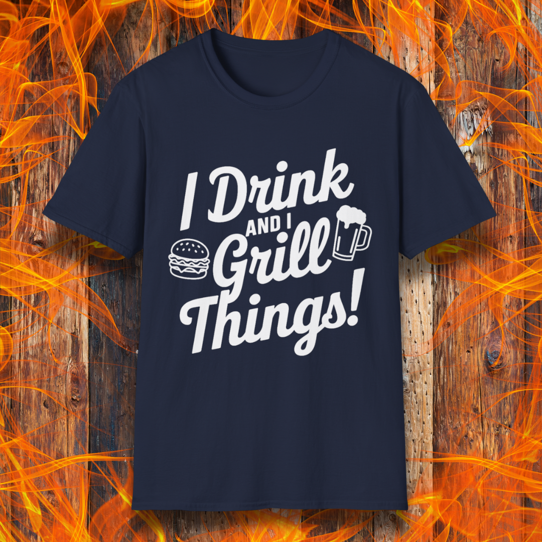 Navy T-shirt featuring a bold, black text design that reads, "I Drink and I Grill Things!" in a playful, script-style font. The design includes small illustrations of a burger and a frothy beer mug, emphasizing a fun and laid-back grilling theme. The background showcases a rustic wooden surface with bright orange flames, enhancing the fiery BBQ aesthetic.