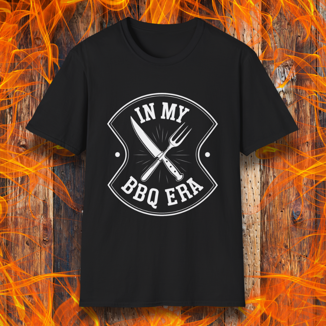 Black T-shirt featuring a bold black design with the phrase "IN MY BBQ ERA" inside a shield-like emblem. At the center of the emblem, there is a crossed knife and grilling fork, symbolizing BBQ culture. The background showcases a rustic wooden texture with bright orange flames, adding a fiery and intense grilling aesthetic.