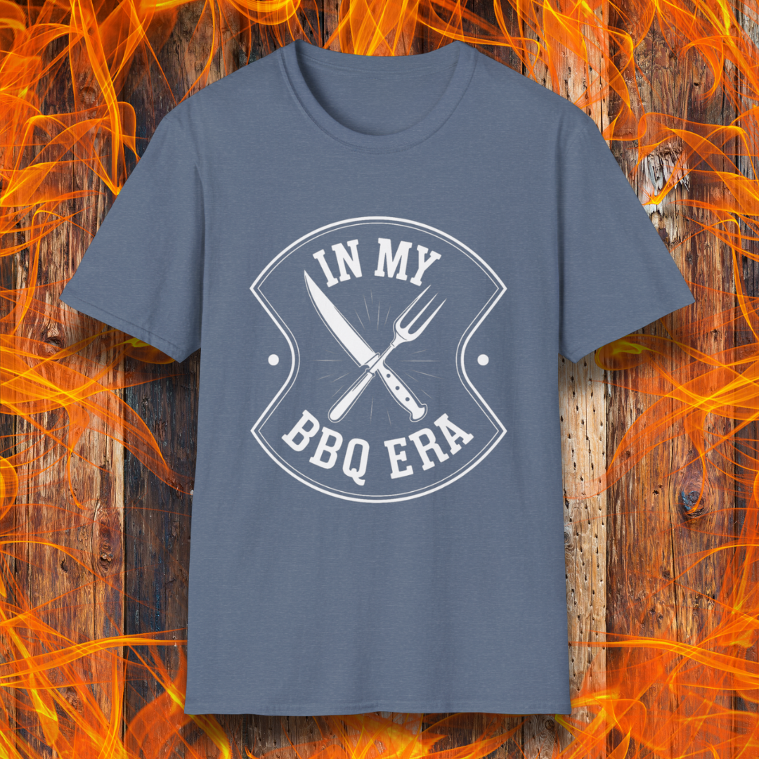 Heather Indigo T-shirt featuring a bold black design with the phrase "IN MY BBQ ERA" inside a shield-like emblem. At the center of the emblem, there is a crossed knife and grilling fork, symbolizing BBQ culture. The background showcases a rustic wooden texture with bright orange flames, adding a fiery and intense grilling aesthetic.
