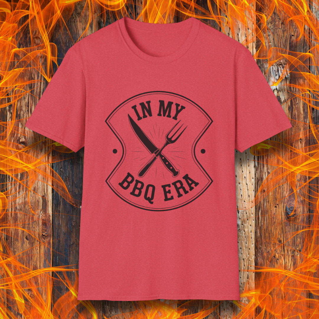 Heather Red T-shirt featuring a bold black design with the phrase "IN MY BBQ ERA" inside a shield-like emblem. At the center of the emblem, there is a crossed knife and grilling fork, symbolizing BBQ culture. The background showcases a rustic wooden texture with bright orange flames, adding a fiery and intense grilling aesthetic.