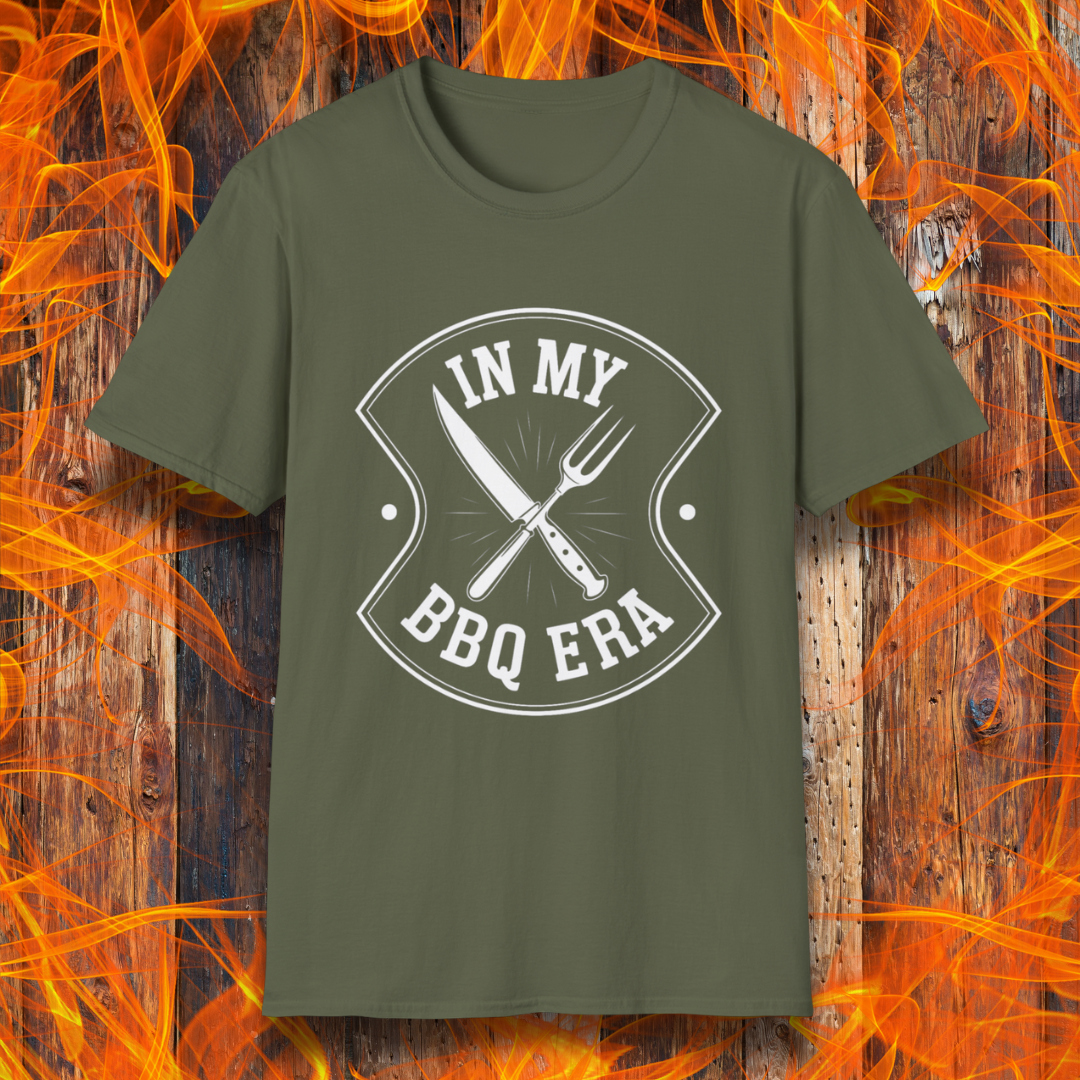 Military Green T-shirt featuring a bold black design with the phrase "IN MY BBQ ERA" inside a shield-like emblem. At the center of the emblem, there is a crossed knife and grilling fork, symbolizing BBQ culture. The background showcases a rustic wooden texture with bright orange flames, adding a fiery and intense grilling aesthetic.
