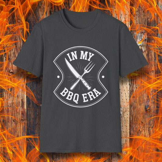 Dark Heather T-shirt featuring a bold black design with the phrase "IN MY BBQ ERA" inside a shield-like emblem. At the center of the emblem, there is a crossed knife and grilling fork, symbolizing BBQ culture. The background showcases a rustic wooden texture with bright orange flames, adding a fiery and intense grilling aesthetic.