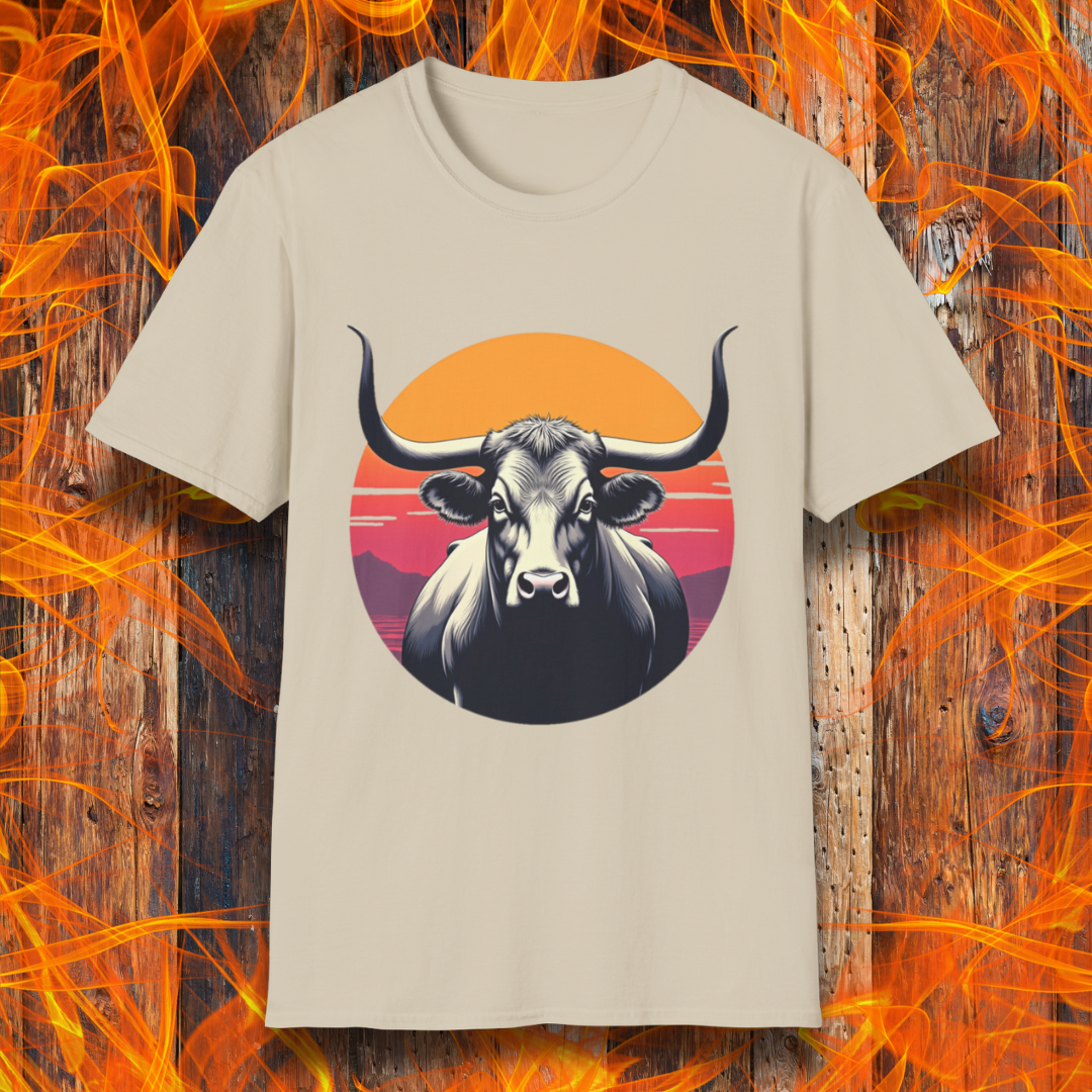 Sand T-shirt featuring a striking design of a longhorn bull staring forward with an intense gaze. The bull is set against a circular backdrop of a sunset with warm orange, red, and pink hues, along with silhouetted mountains in the background. The design conveys strength, confidence, and a bold Western aesthetic, appealing to BBQ and cattle enthusiasts.