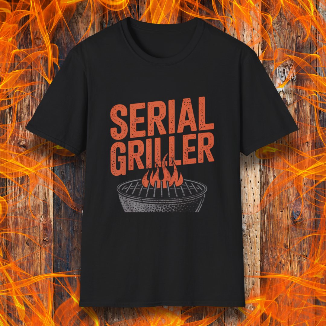 Black T-shirt featuring a bold, red-orange text design that reads "SERIAL GRILLER." Below the text, there's a black and white illustration of a round charcoal grill with flames rising from the grates. The background consists of a rustic wooden surface with bright orange flames surrounding the shirt, creating a fiery grilling-themed aesthetic.