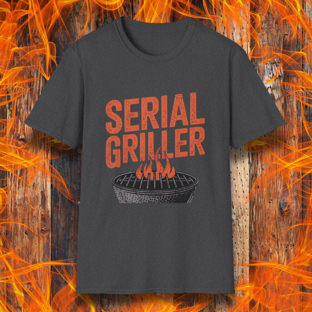 Dark Heather T-shirt featuring a bold, red-orange text design that reads "SERIAL GRILLER." Below the text, there's a black and white illustration of a round charcoal grill with flames rising from the grates. The background consists of a rustic wooden surface with bright orange flames surrounding the shirt, creating a fiery grilling-themed aesthetic.