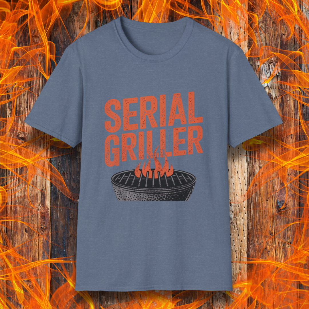 Heather Indigo T-shirt featuring a bold, red-orange text design that reads "SERIAL GRILLER." Below the text, there's a black and white illustration of a round charcoal grill with flames rising from the grates. The background consists of a rustic wooden surface with bright orange flames surrounding the shirt, creating a fiery grilling-themed aesthetic.