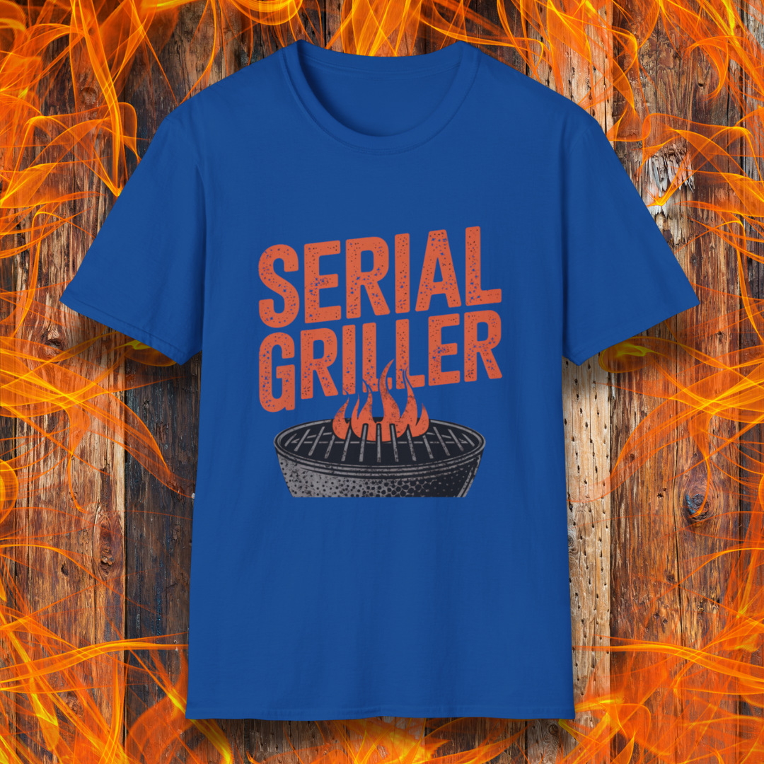 Royal Blue T-shirt featuring a bold, red-orange text design that reads "SERIAL GRILLER." Below the text, there's a black and white illustration of a round charcoal grill with flames rising from the grates. The background consists of a rustic wooden surface with bright orange flames surrounding the shirt, creating a fiery grilling-themed aesthetic.