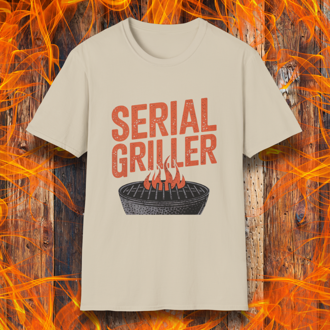 Sand T-shirt featuring a bold, red-orange text design that reads "SERIAL GRILLER." Below the text, there's a black and white illustration of a round charcoal grill with flames rising from the grates. The background consists of a rustic wooden surface with bright orange flames surrounding the shirt, creating a fiery grilling-themed aesthetic.