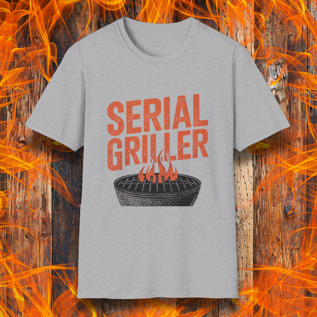 Sport Grey T-shirt featuring a bold, red-orange text design that reads "SERIAL GRILLER." Below the text, there's a black and white illustration of a round charcoal grill with flames rising from the grates. The background consists of a rustic wooden surface with bright orange flames surrounding the shirt, creating a fiery grilling-themed aesthetic.