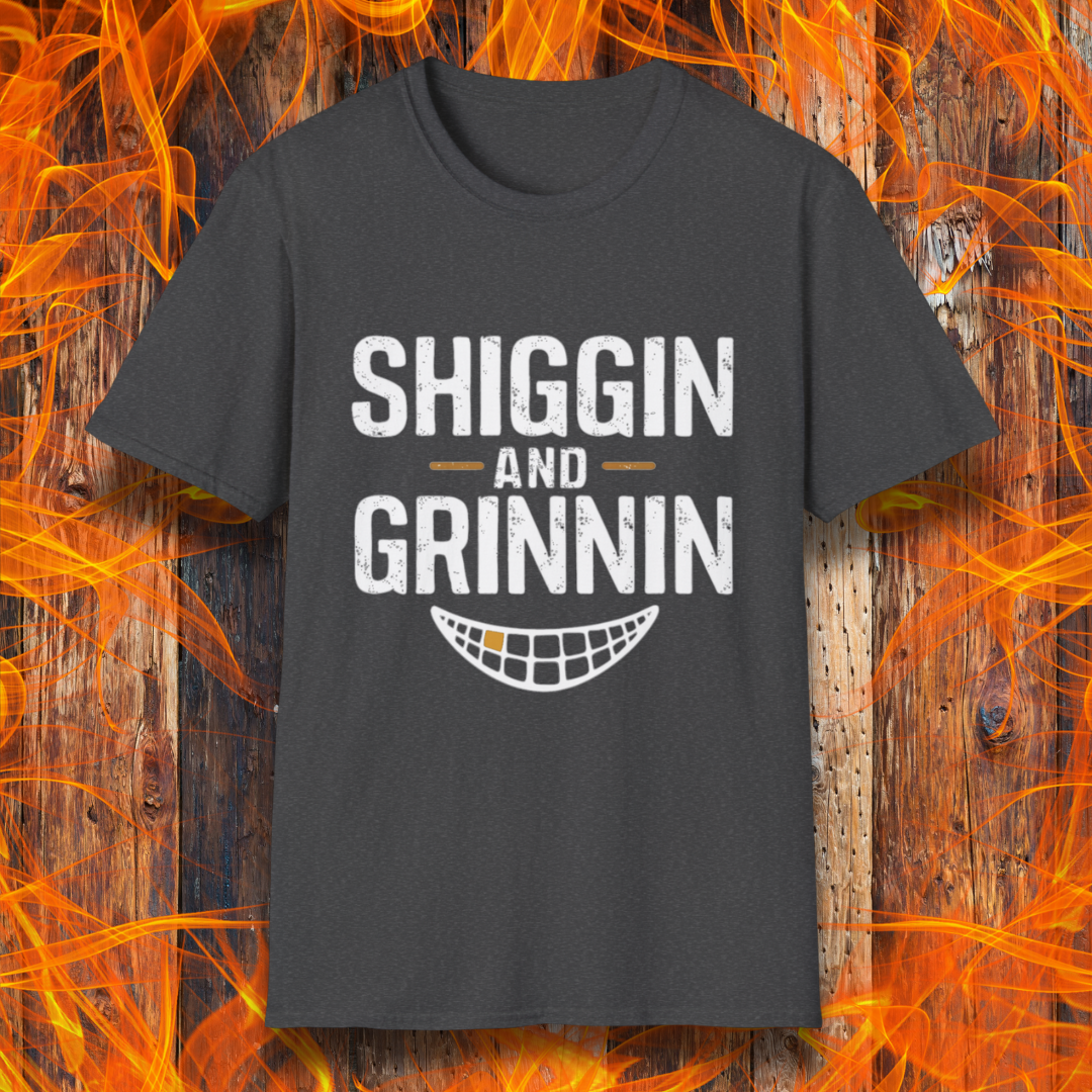 Dark Heather T-shirt featuring the phrase 'Shiggin and Grinnin' in bold, distressed white lettering. Below the text is a stylized smiling grill grate graphic. The design is set against a rustic wooden background with flames surrounding the edges, giving it a fiery barbecue-themed look.