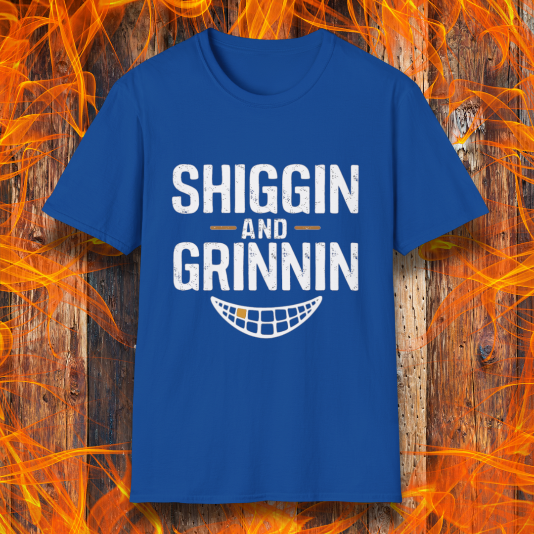 Royal Blue T-shirt featuring the phrase 'Shiggin and Grinnin' in bold, distressed white lettering. Below the text is a stylized smiling grill grate graphic. The design is set against a rustic wooden background with flames surrounding the edges, giving it a fiery barbecue-themed look.