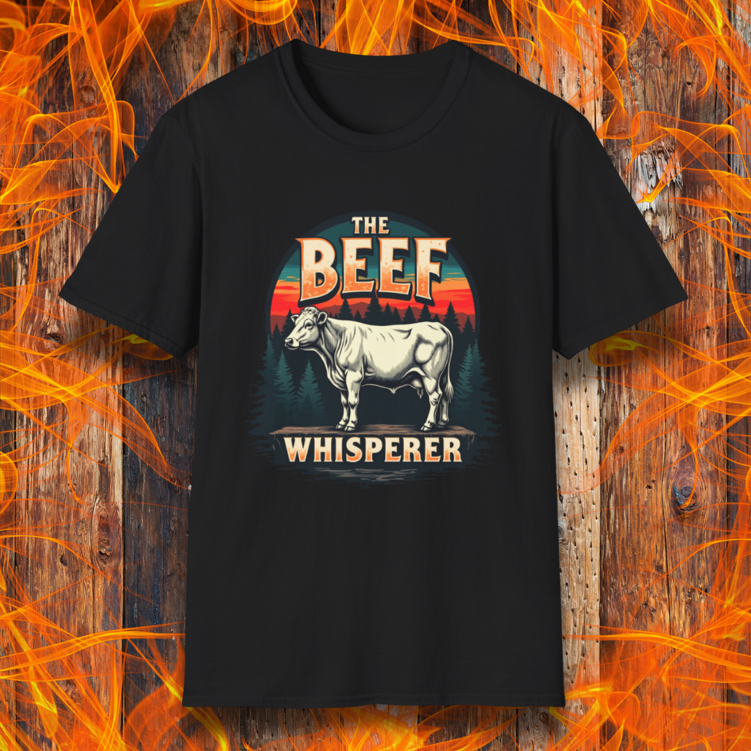 Black T-shirt featuring a bold design with the words "The Beef Whisperer" in a vintage western-style font. Above the text, a detailed illustration of a cow stands on a patch of ground, framed by a scenic forest and a sunset with deep orange and red hues. The design has a rustic, outdoorsy feel, appealing to BBQ lovers and beef enthusiasts.