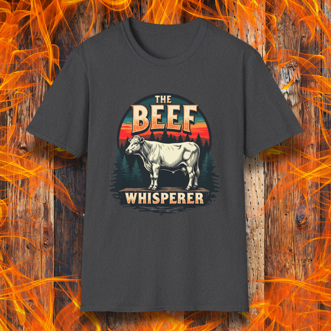 Dark Heather  T-shirt featuring a bold design with the words "The Beef Whisperer" in a vintage western-style font. Above the text, a detailed illustration of a cow stands on a patch of ground, framed by a scenic forest and a sunset with deep orange and red hues. The design has a rustic, outdoorsy feel, appealing to BBQ lovers and beef enthusiasts.