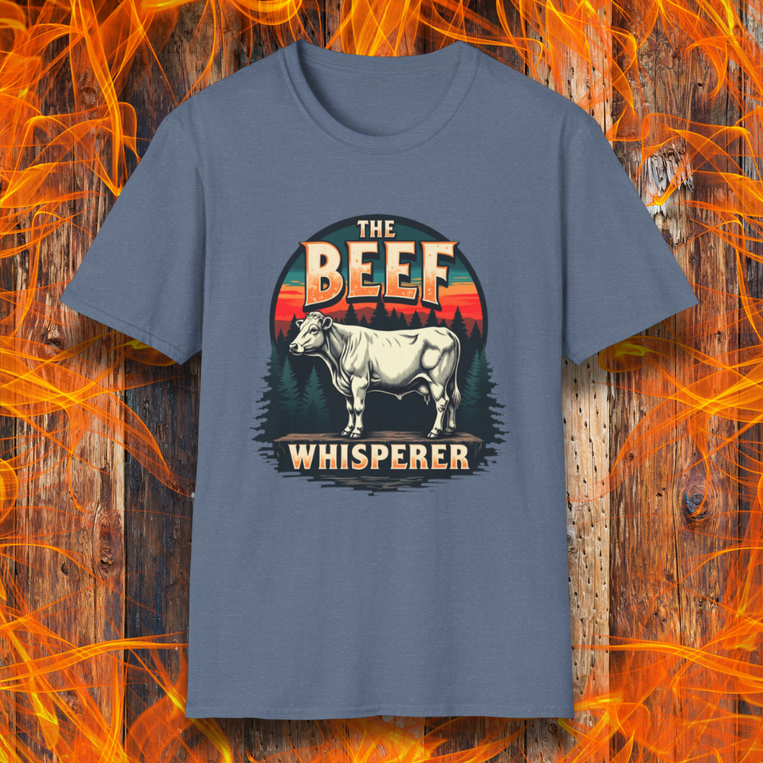 Heather Indigo T-shirt featuring a bold design with the words "The Beef Whisperer" in a vintage western-style font. Above the text, a detailed illustration of a cow stands on a patch of ground, framed by a scenic forest and a sunset with deep orange and red hues. The design has a rustic, outdoorsy feel, appealing to BBQ lovers and beef enthusiasts.