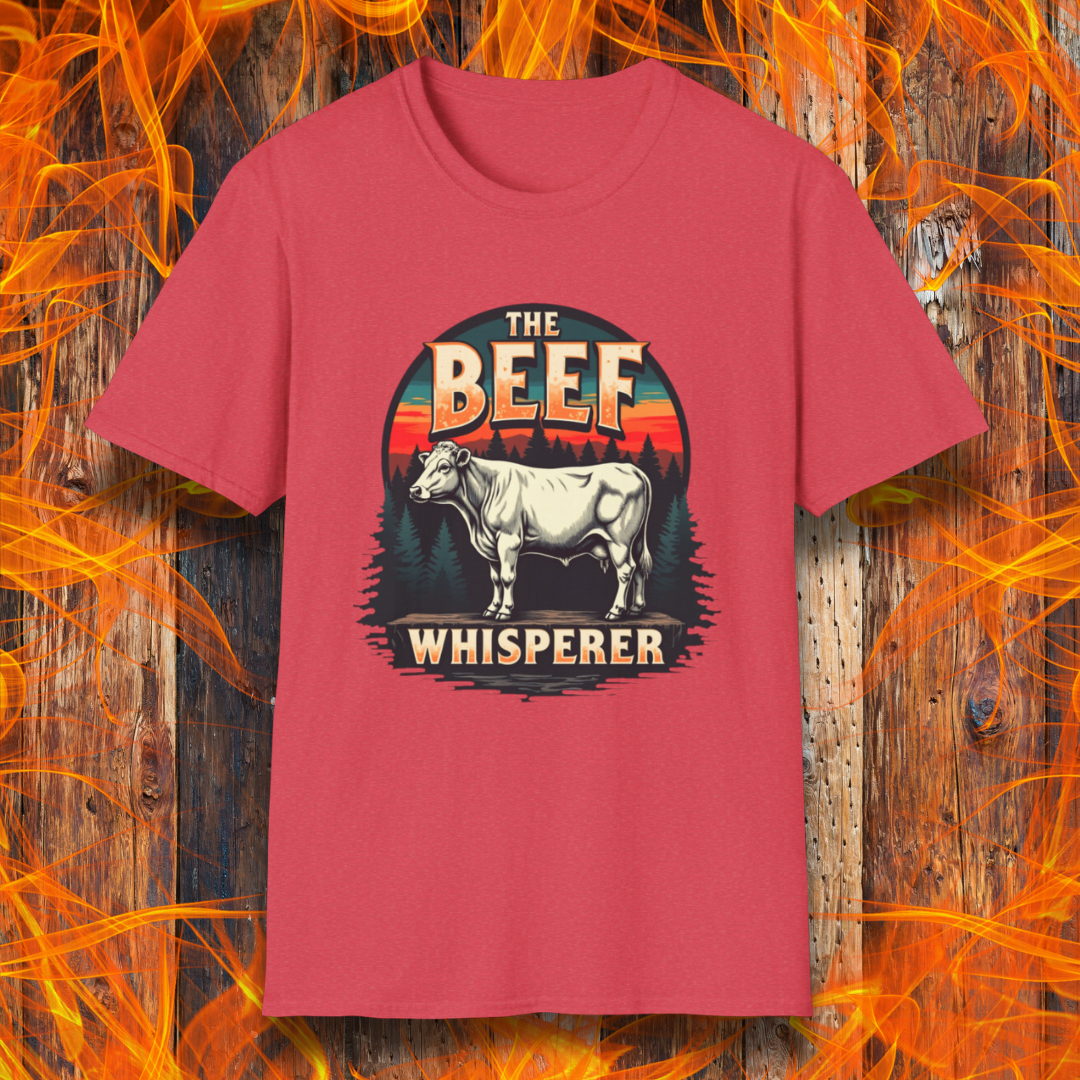 Heather Red  T-shirt featuring a bold design with the words "The Beef Whisperer" in a vintage western-style font. Above the text, a detailed illustration of a cow stands on a patch of ground, framed by a scenic forest and a sunset with deep orange and red hues. The design has a rustic, outdoorsy feel, appealing to BBQ lovers and beef enthusiasts.