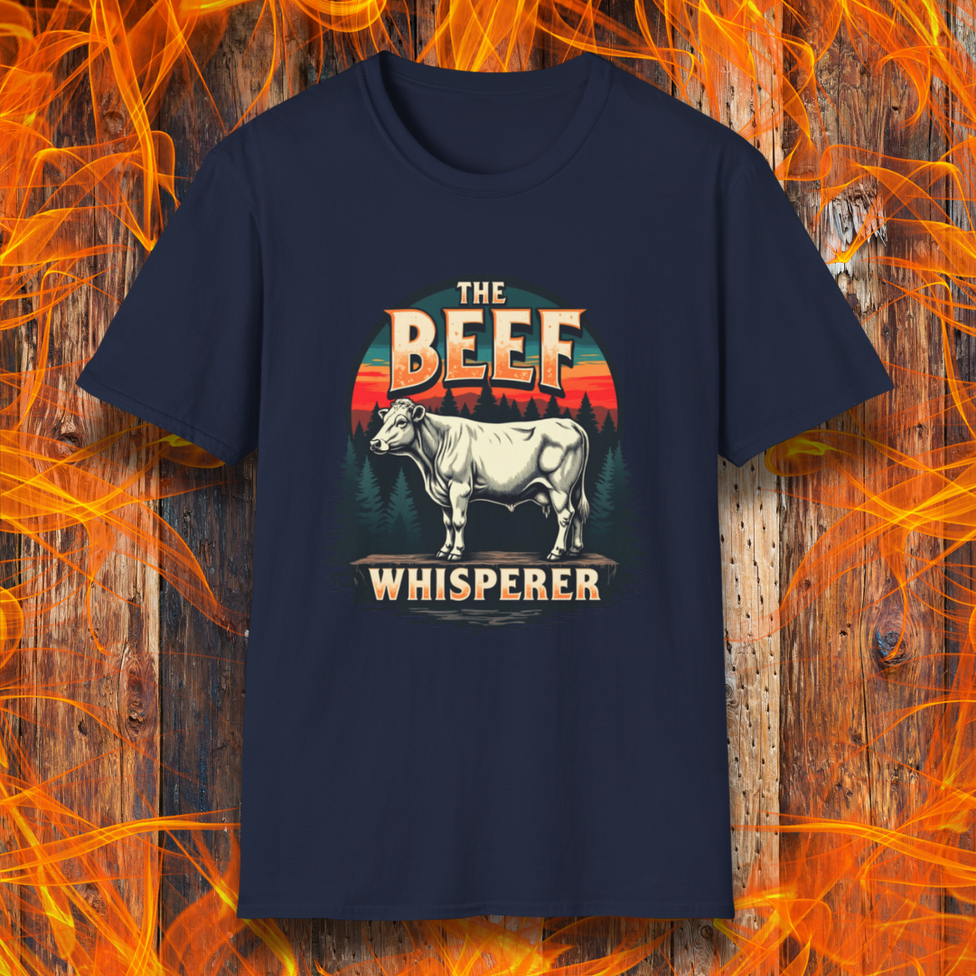 Navy T-shirt featuring a bold design with the words "The Beef Whisperer" in a vintage western-style font. Above the text, a detailed illustration of a cow stands on a patch of ground, framed by a scenic forest and a sunset with deep orange and red hues. The design has a rustic, outdoorsy feel, appealing to BBQ lovers and beef enthusiasts.