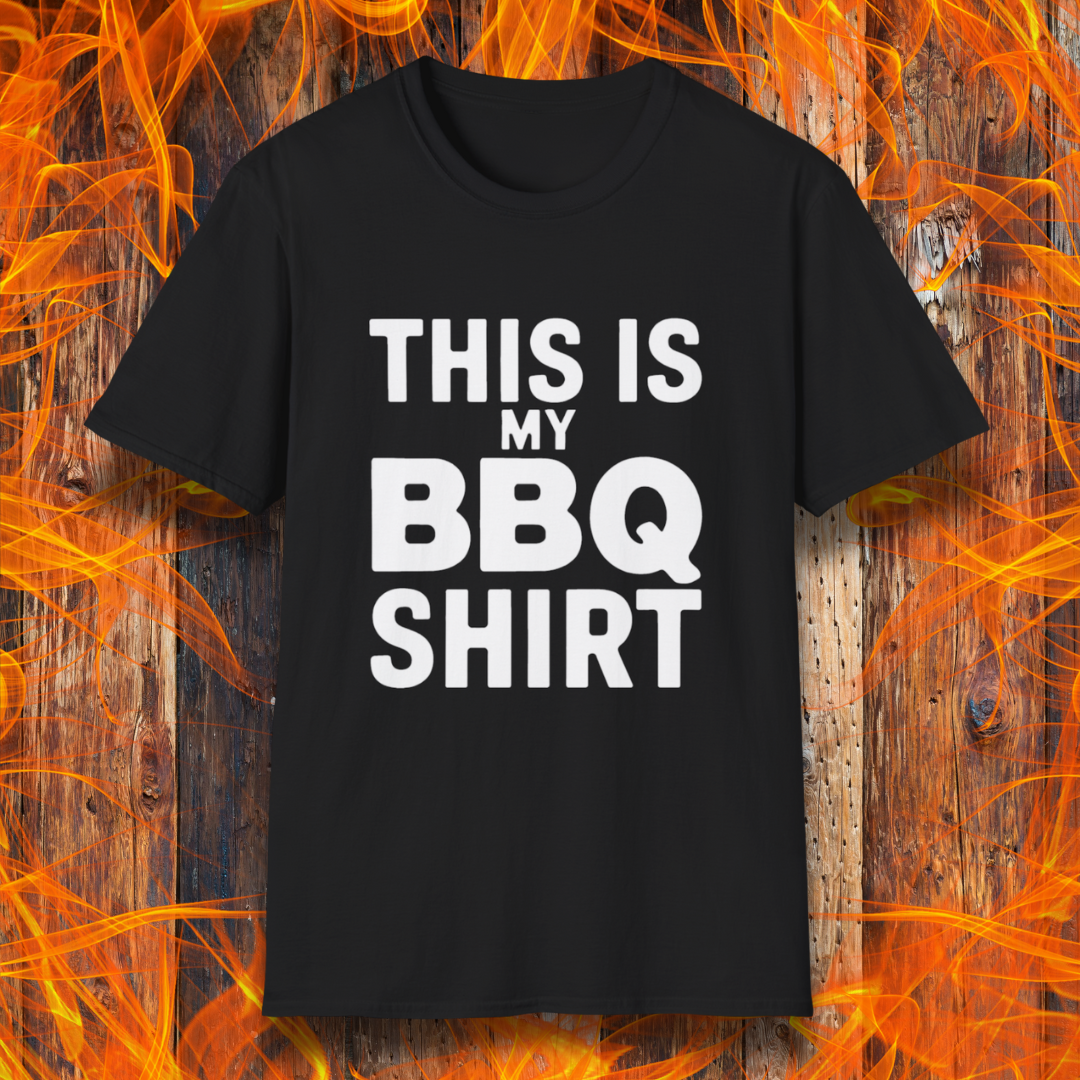 Black T-shirt with bold white text that reads "THIS IS MY BBQ SHIRT." The shirt is displayed against a fiery wooden background, reinforcing its BBQ and grilling theme.