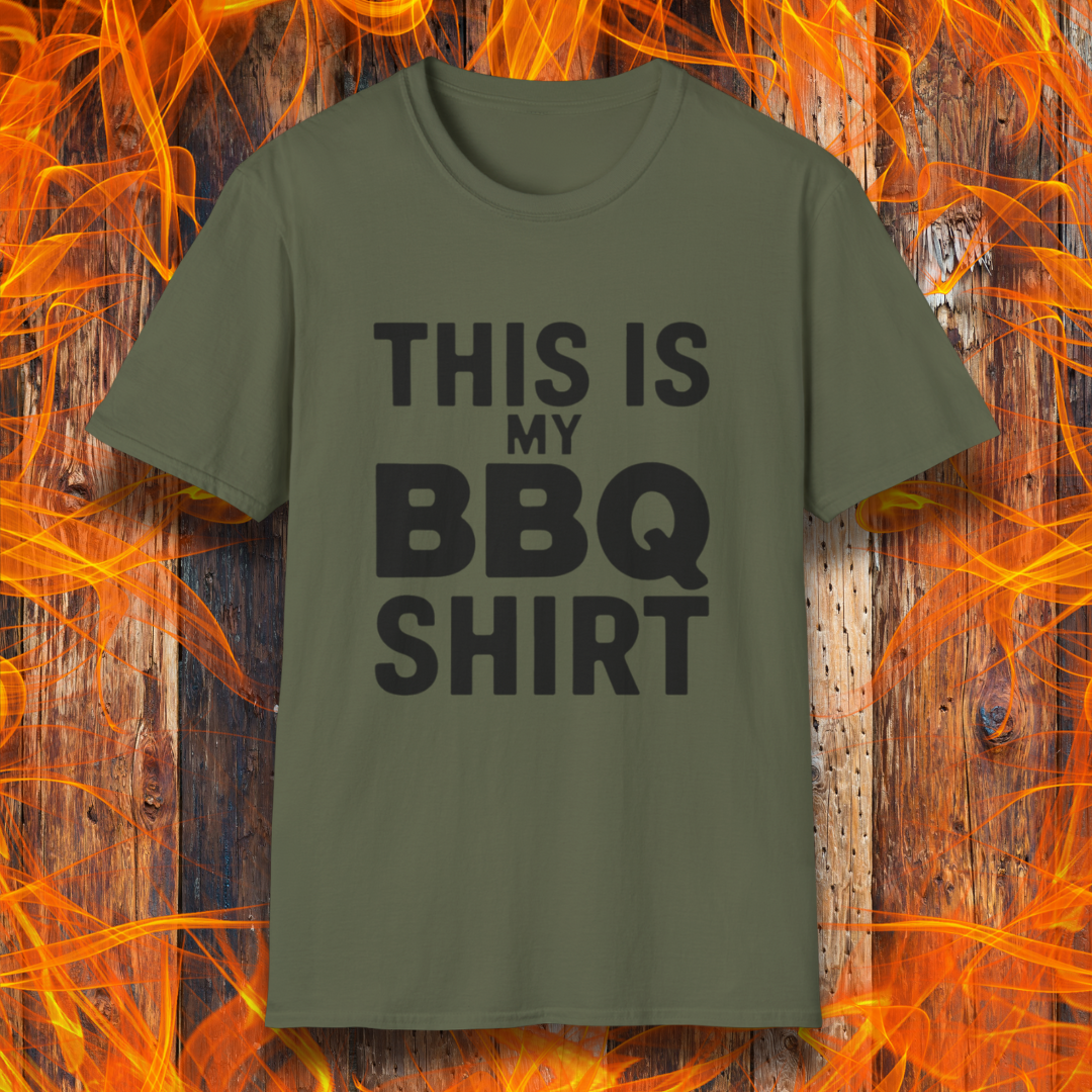 Military Green T-shirt with bold Black text that reads "THIS IS MY BBQ SHIRT." The shirt is displayed against a fiery wooden background, reinforcing its BBQ and grilling theme.