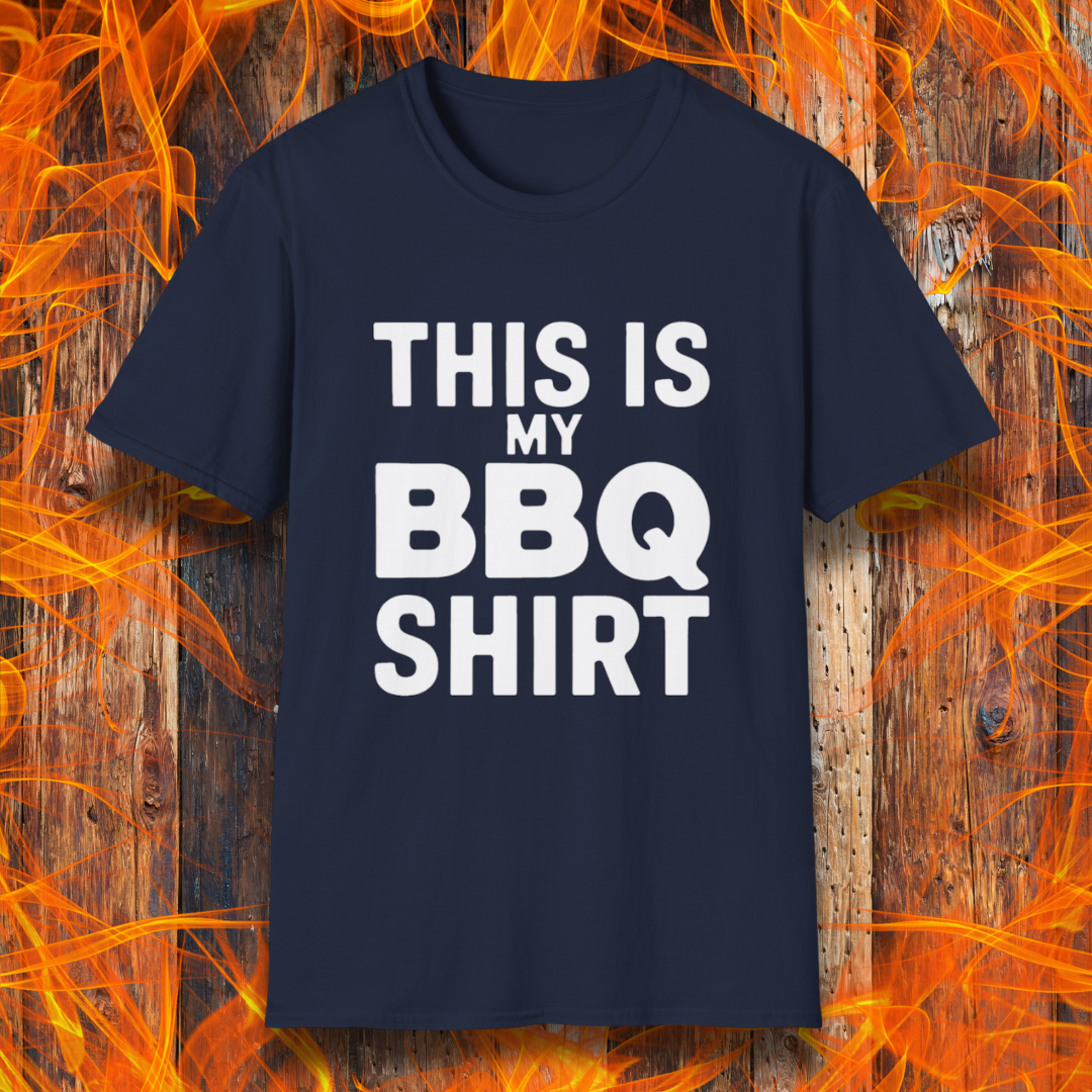 Navy T-shirt with bold white text that reads "THIS IS MY BBQ SHIRT." The shirt is displayed against a fiery wooden background, reinforcing its BBQ and grilling theme.