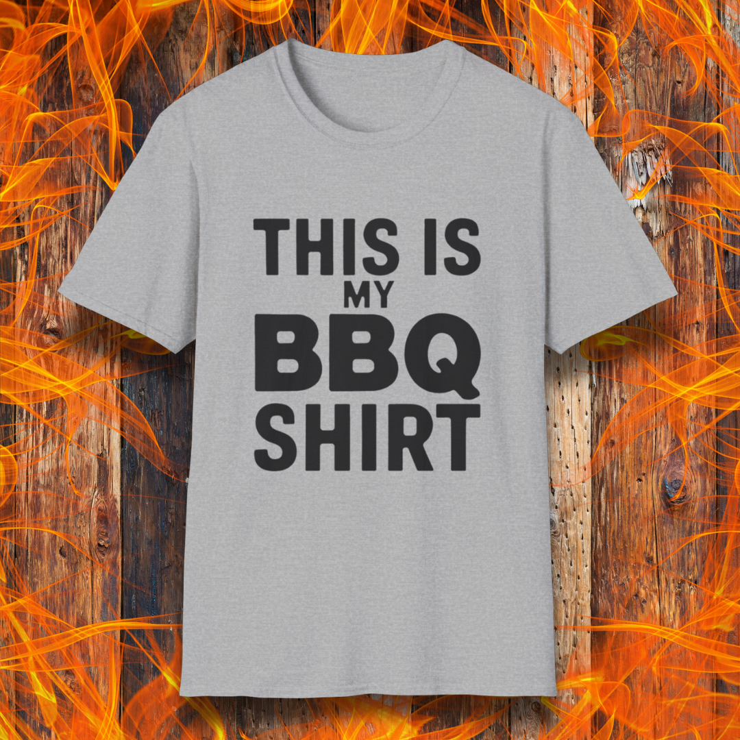 Sport Grey T-shirt with bold black text that reads "THIS IS MY BBQ SHIRT." The shirt is displayed against a fiery wooden background, reinforcing its BBQ and grilling theme.