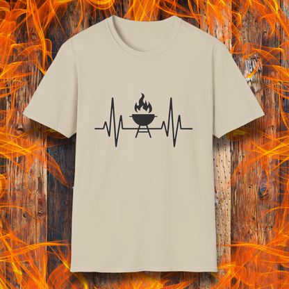 Sand colored T-shirt featuring a minimalist black design of a heartbeat line that transitions into a flaming charcoal grill in the center. The design symbolizes a passion for BBQ, integrating a clean and modern aesthetic. The background consists of a rustic wooden surface with vivid orange flames, enhancing the fiery and grilling-focused theme.