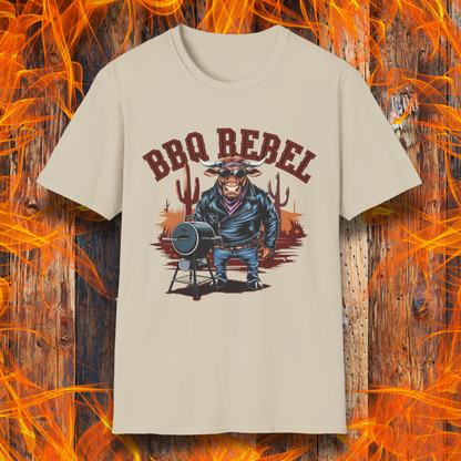 Sand T-shirt featuring a bold and rebellious BBQ-themed design. The centerpiece is an anthropomorphic bull dressed like a biker, wearing a black leather jacket, blue jeans, and a red bandana, exuding a tough and confident attitude. He stands next to a classic barrel-style smoker. The background includes red flames and grill forks, adding to the edgy, fiery vibe. Above the character, "BBQ REBEL" is displayed in bold, vintage-style red lettering.  
