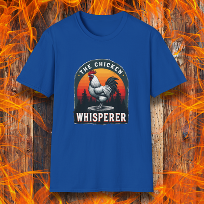 Royal Blue T-shirt featuring a bold design with a proud rooster standing on a wooden stump. The rooster is framed by an arched emblem with a scenic sunset gradient in orange, red, and yellow hues, along with a silhouette of pine trees in the background. The text "THE CHICKEN WHISPERER" is displayed prominently, giving the design a humorous and rustic feel, perfect for backyard BBQ enthusiasts and poultry lovers.