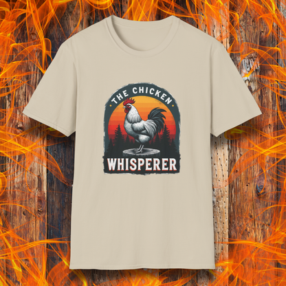 Sand T-shirt featuring a bold design with a proud rooster standing on a wooden stump. The rooster is framed by an arched emblem with a scenic sunset gradient in orange, red, and yellow hues, along with a silhouette of pine trees in the background. The text "THE CHICKEN WHISPERER" is displayed prominently, giving the design a humorous and rustic feel, perfect for backyard BBQ enthusiasts and poultry lovers.