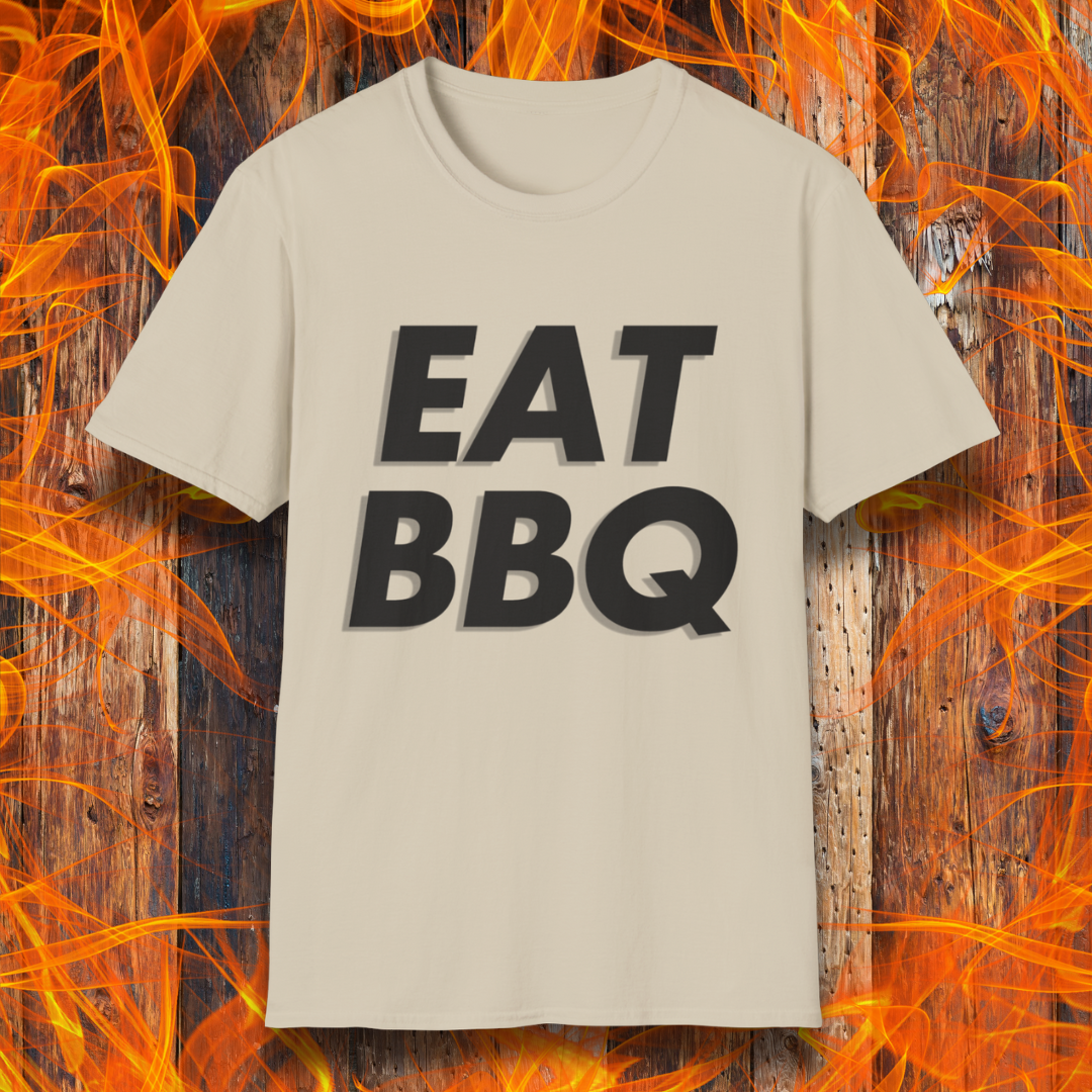 Sand T-shirt featuring bold, blocky white text that reads "EAT BBQ" with a 3D effect. The shirt is displayed against a fiery wooden background, emphasizing the BBQ theme.