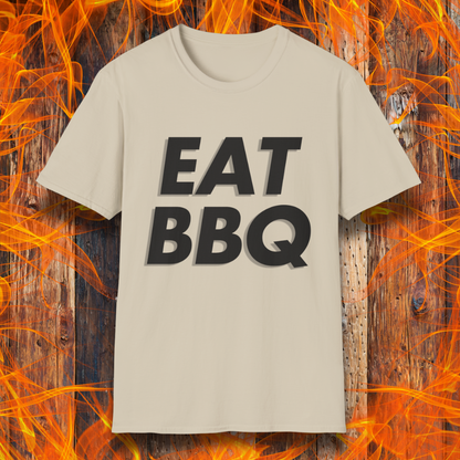 Sand T-shirt featuring bold, blocky white text that reads "EAT BBQ" with a 3D effect. The shirt is displayed against a fiery wooden background, emphasizing the BBQ theme.