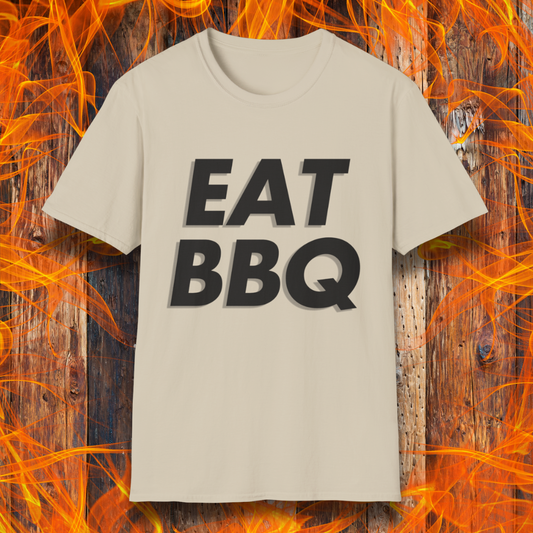 Sand T-shirt featuring bold, blocky white text that reads "EAT BBQ" with a 3D effect. The shirt is displayed against a fiery wooden background, emphasizing the BBQ theme.