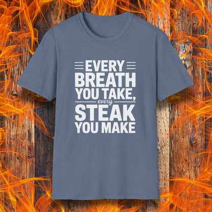 Heather Indigo T-shirt with bold white text that reads "Every Breath You Take, Every Steak You Make," displayed over a rustic wood and flame background — a humorous grilling shirt perfect for steak fans.