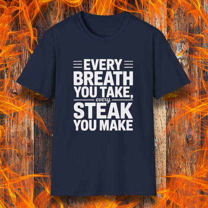 Navy T-shirt with bold white text that reads "Every Breath You Take, Every Steak You Make," displayed over a rustic wood and flame background — a humorous grilling shirt perfect for steak fans.