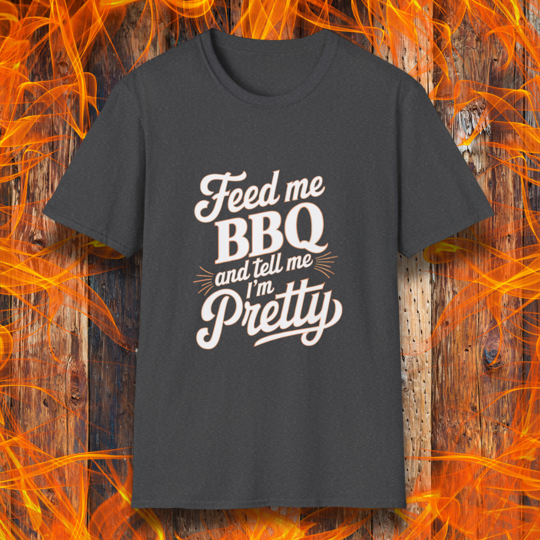 Dark Heather T-shirt featuring a playful and bold typographic design that reads, "Feed me BBQ and tell me I'm Pretty." The text is styled in a mix of script and block fonts, with decorative accents emphasizing "BBQ" and "Pretty." The shirt is set against a rustic wooden background with vibrant orange flames, reinforcing the fiery BBQ theme.