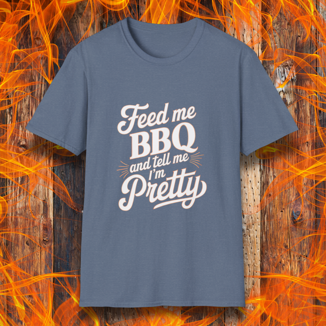 Heather Indigo T-shirt featuring a playful and bold typographic design that reads, "Feed me BBQ and tell me I'm Pretty." The text is styled in a mix of script and block fonts, with decorative accents emphasizing "BBQ" and "Pretty." The shirt is set against a rustic wooden background with vibrant orange flames, reinforcing the fiery BBQ theme.