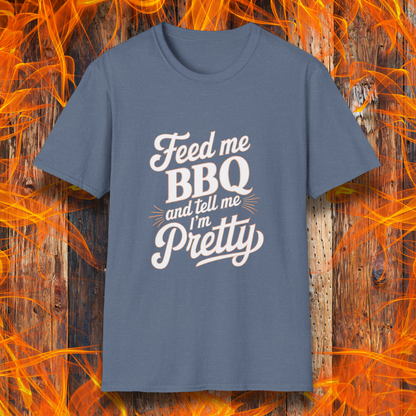 Heather Indigo T-shirt featuring a playful and bold typographic design that reads, "Feed me BBQ and tell me I'm Pretty." The text is styled in a mix of script and block fonts, with decorative accents emphasizing "BBQ" and "Pretty." The shirt is set against a rustic wooden background with vibrant orange flames, reinforcing the fiery BBQ theme.