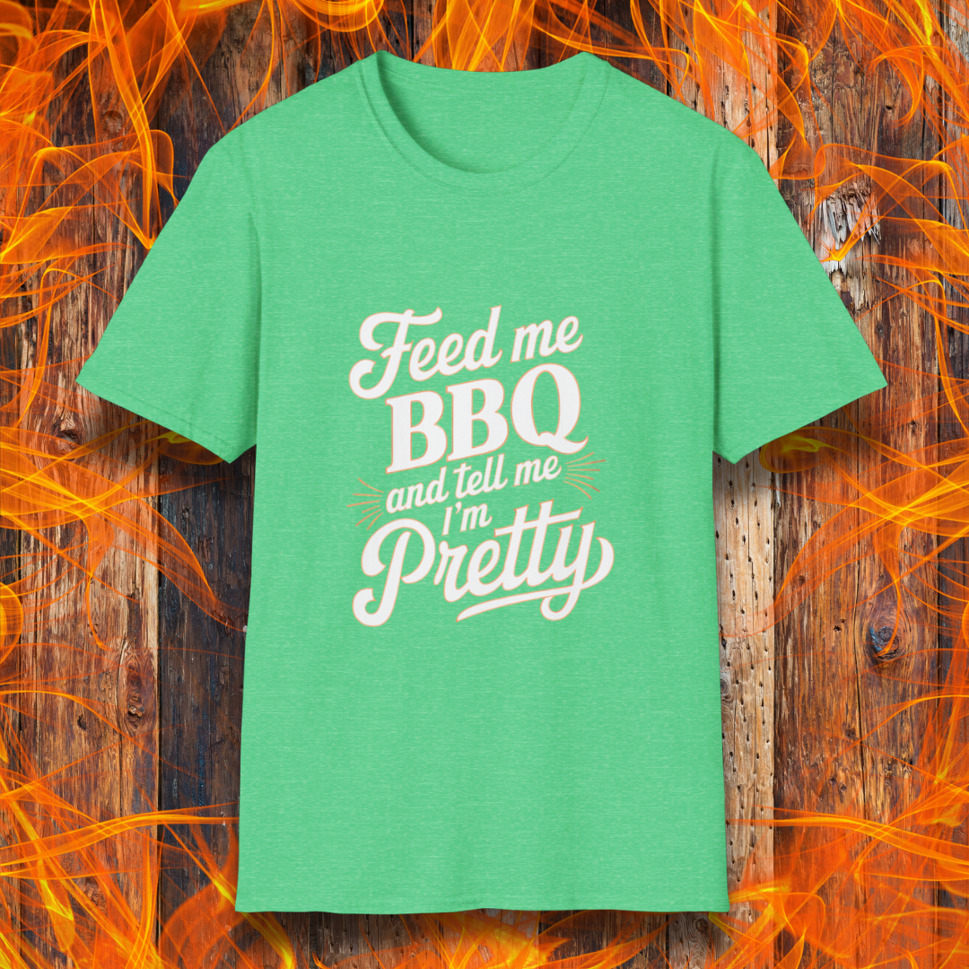 Heather Irish Green T-shirt featuring a playful and bold typographic design that reads, "Feed me BBQ and tell me I'm Pretty." The text is styled in a mix of script and block fonts, with decorative accents emphasizing "BBQ" and "Pretty." The shirt is set against a rustic wooden background with vibrant orange flames, reinforcing the fiery BBQ theme.