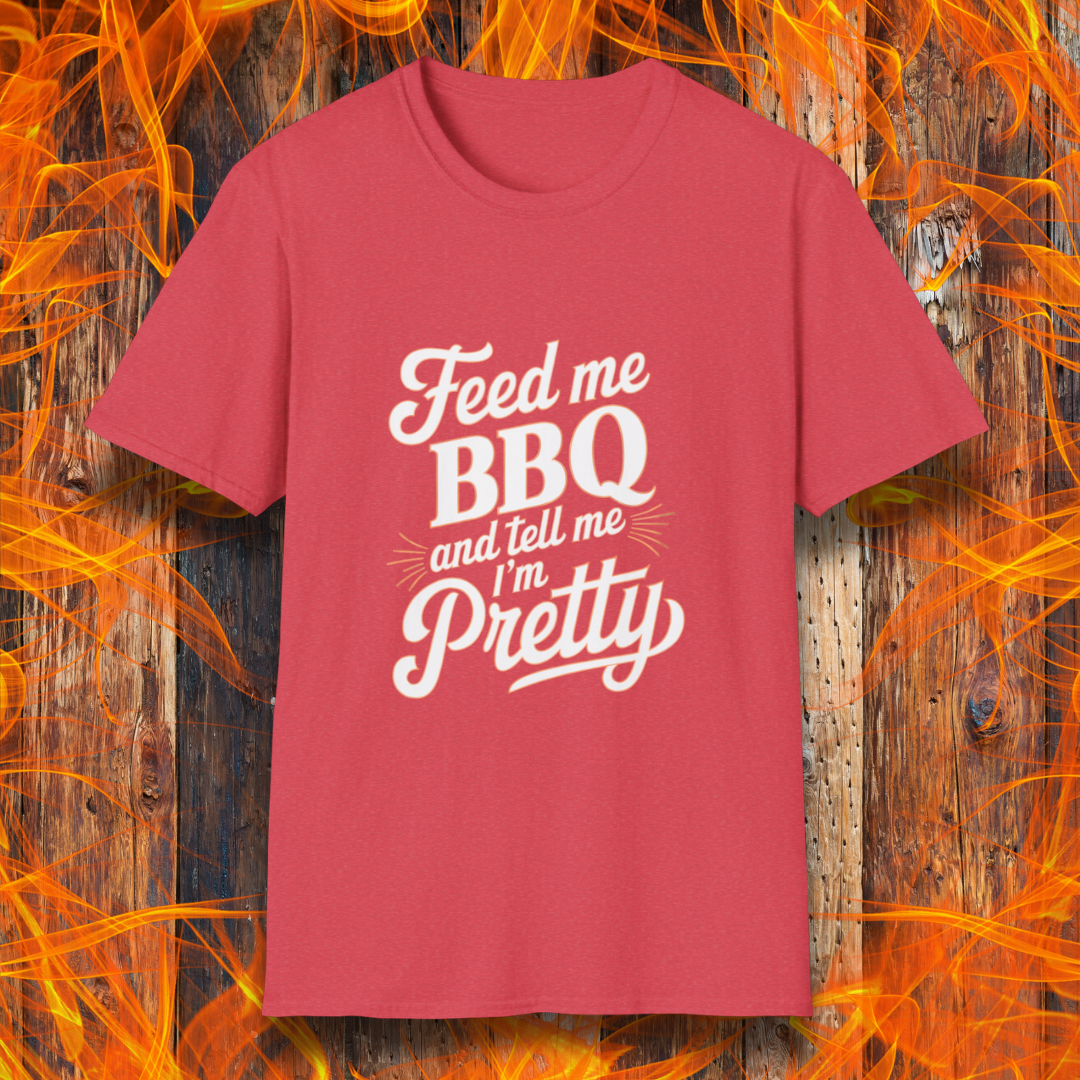 Heather Red T-shirt featuring a playful and bold typographic design that reads, "Feed me BBQ and tell me I'm Pretty." The text is styled in a mix of script and block fonts, with decorative accents emphasizing "BBQ" and "Pretty." The shirt is set against a rustic wooden background with vibrant orange flames, reinforcing the fiery BBQ theme.