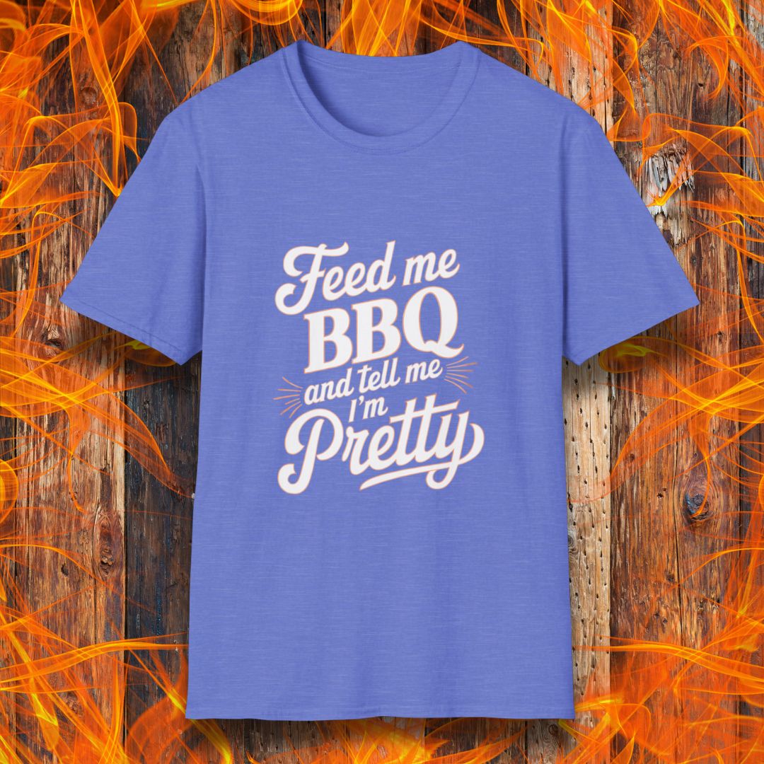 Heather Roayl T-shirt featuring a playful and bold typographic design that reads, "Feed me BBQ and tell me I'm Pretty." The text is styled in a mix of script and block fonts, with decorative accents emphasizing "BBQ" and "Pretty." The shirt is set against a rustic wooden background with vibrant orange flames, reinforcing the fiery BBQ theme.